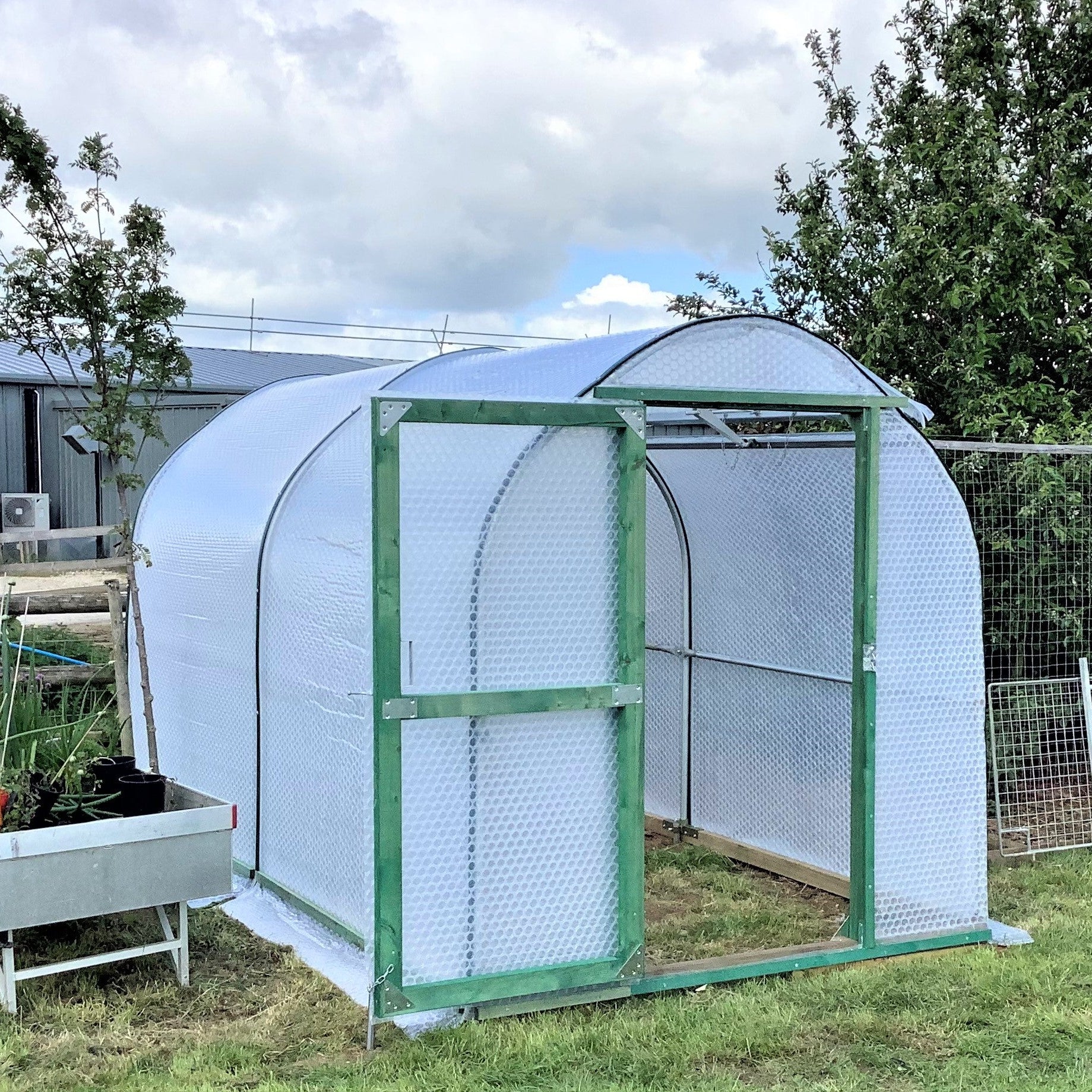 Greenhouse with a wire mesh door on a grassy area with trees and buildings in the background.