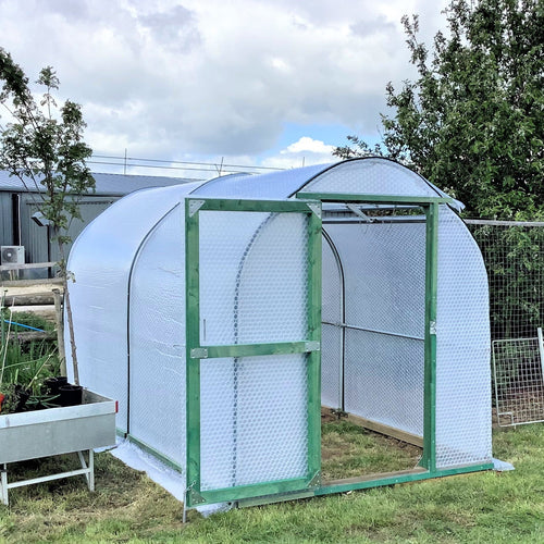 Greenhouse with a wire mesh door on a grassy area with trees and buildings in the background.